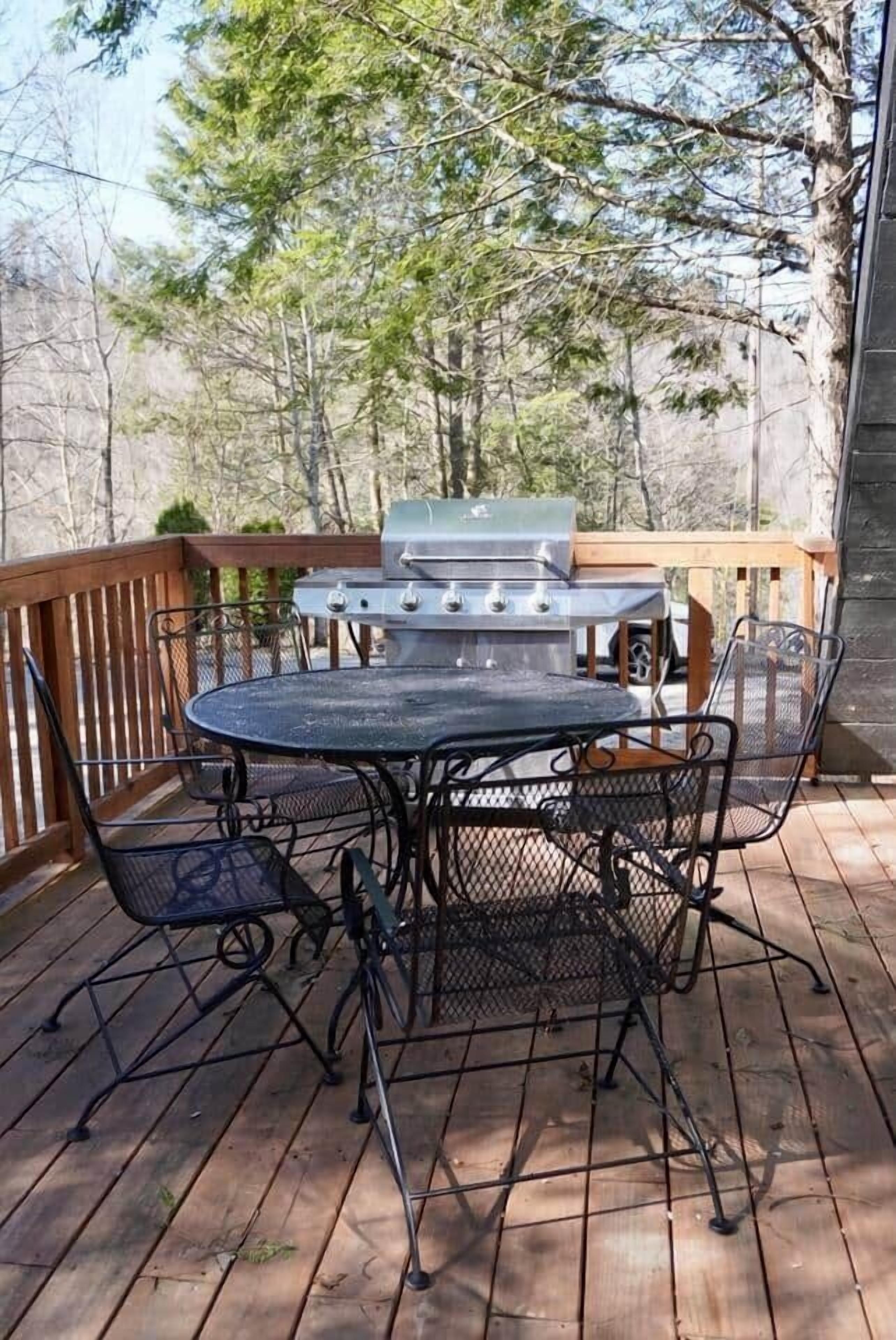 A covered outdoor deck with a round metal table and four chairs, a barbecue grill, and a wooden railing.