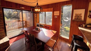 Ein Holzblock-Interieur mit Esstisch, Stühlen, einer Chandelieber und großen Fenstern, die einen Ausblick ins Freie bieten.