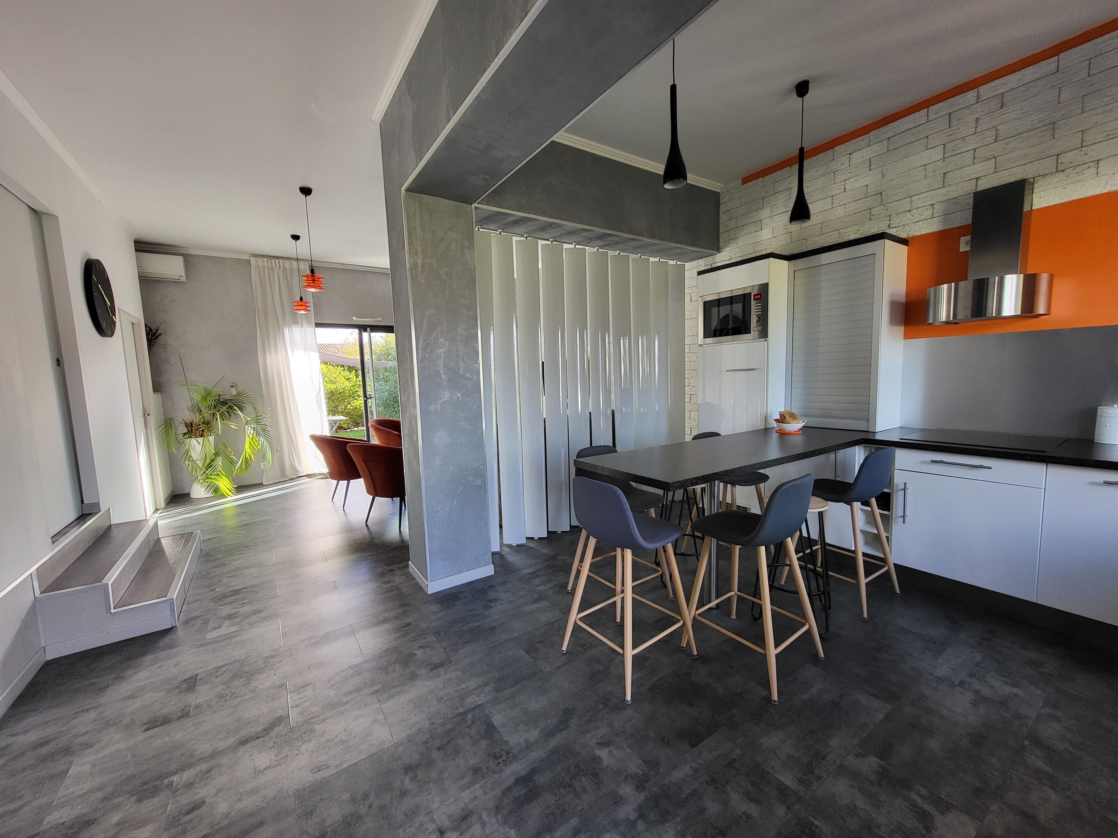 Une cuisine moderne avec un plan de travail noir, des armoires blanches et une hotte de cuisinière en acier inoxydable. On y trouve quatre tabourets de bar et une grande fenêtre donnant sur un paysage verdoyant.
