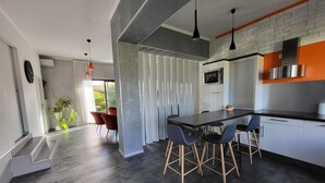 Een moderne keuken met een zwart aanrecht, witte kasten en een rvs afzuigkap. Er zijn vier barkrukken en een groot raam met uitzicht op groen buiten.
