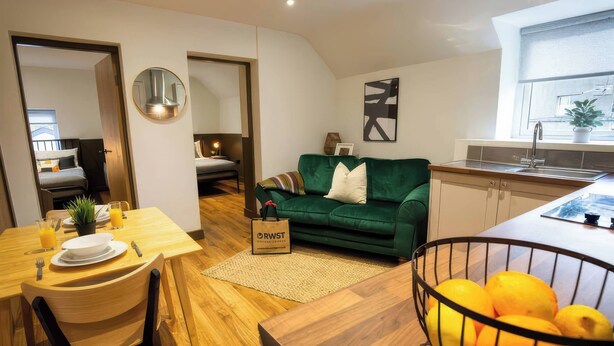 Ein modernes Wohnzimmer mit einem grünen Sofa, einem Esstisch mit Frühstücksgebäck und einer Küchenzeile mit einer Obstschale.