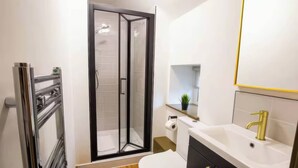 Ein modernes Badezimmer mit Duschkabine aus Glas, Toilette, Waschbecken mit Goldarmatur und einer kleinen Ablage mit Pflanze.
