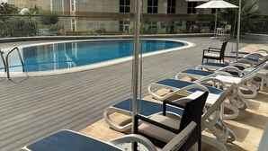 Uma área à beira da piscina com espreguiçadeiras e guarda-sóis, uma piscina e um edifício ao fundo.