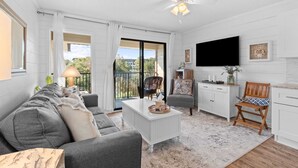 A modern living room with a white sofa, a coffee table, and a TV. There is a balcony with a view of greenery. - Salty Smiles (Hilton Head Island)