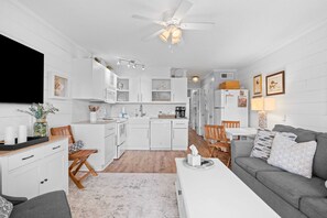 A modern kitchen with white cabinetry, a dining area with a grey sofa, and a ceiling fan. - Salty Smiles (Hilton Head Island)