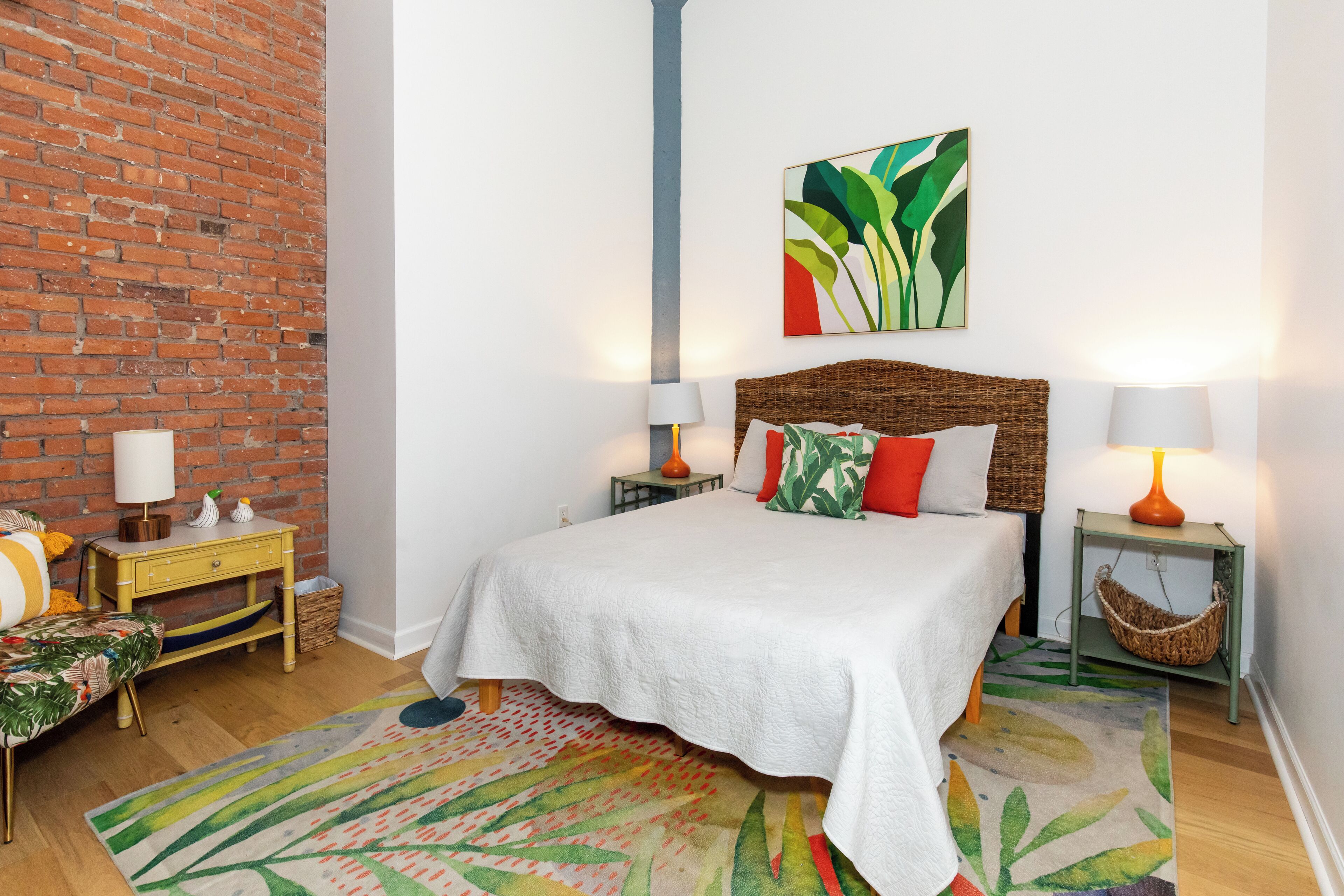 Une chambre avec un mur en briques, un grand lit avec une literie blanche, deux tables de chevet avec des lampes, un tapis coloré et un tableau abstrait accroché au mur.