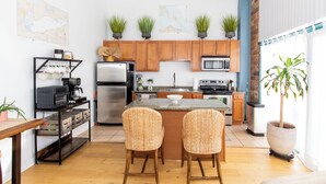 Una cocina moderna con armarios de madera, un refrigerador de acero inoxidable, un microondas y una cafetera. Hay dos sillas de mimbre en una pequeña isla, y plantas en macetas sobre las encimeras.