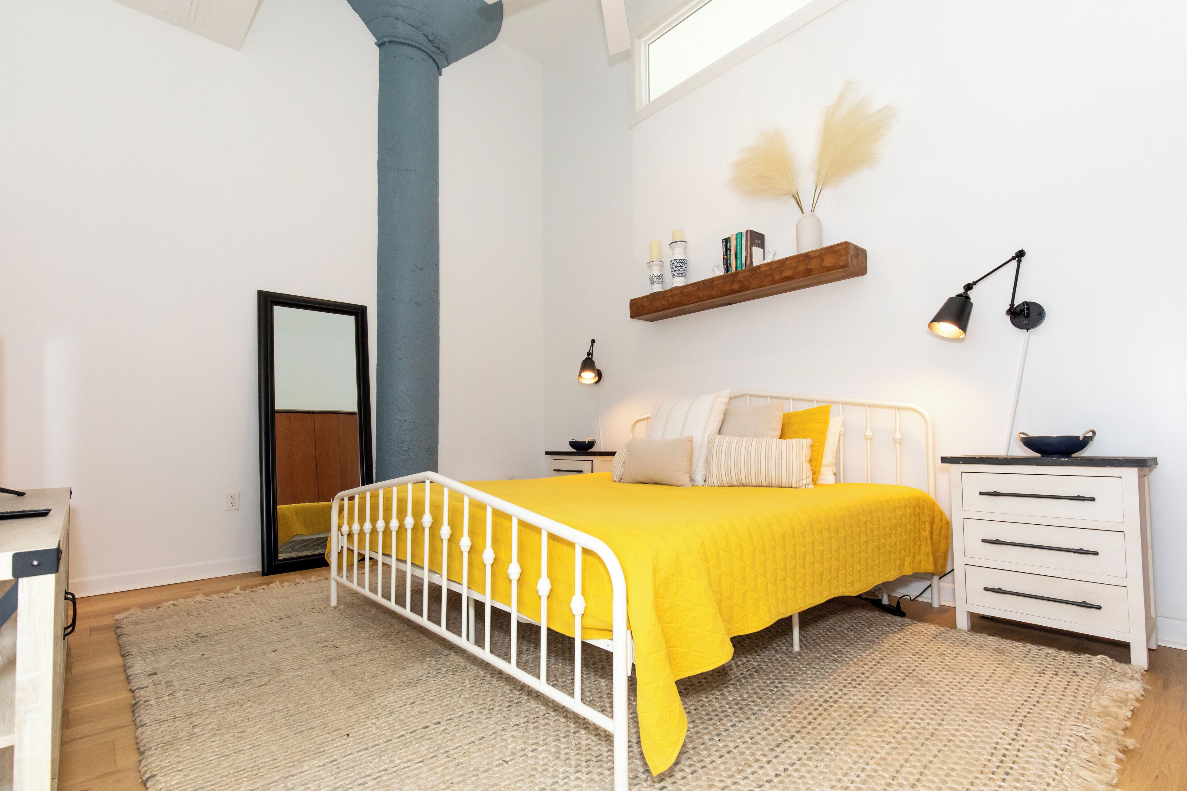 Une chambre à coucher comprenant un lit, des tables de chevet, un miroir, une étagère avec des objets décoratifs et une fenêtre.