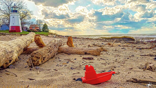 Una playa con madera a la deriva, un juguete rojo y un faro al fondo.