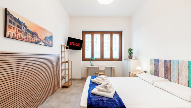Quarto de hotel com cama, escrivaninha, cadeira, televisão e janela.