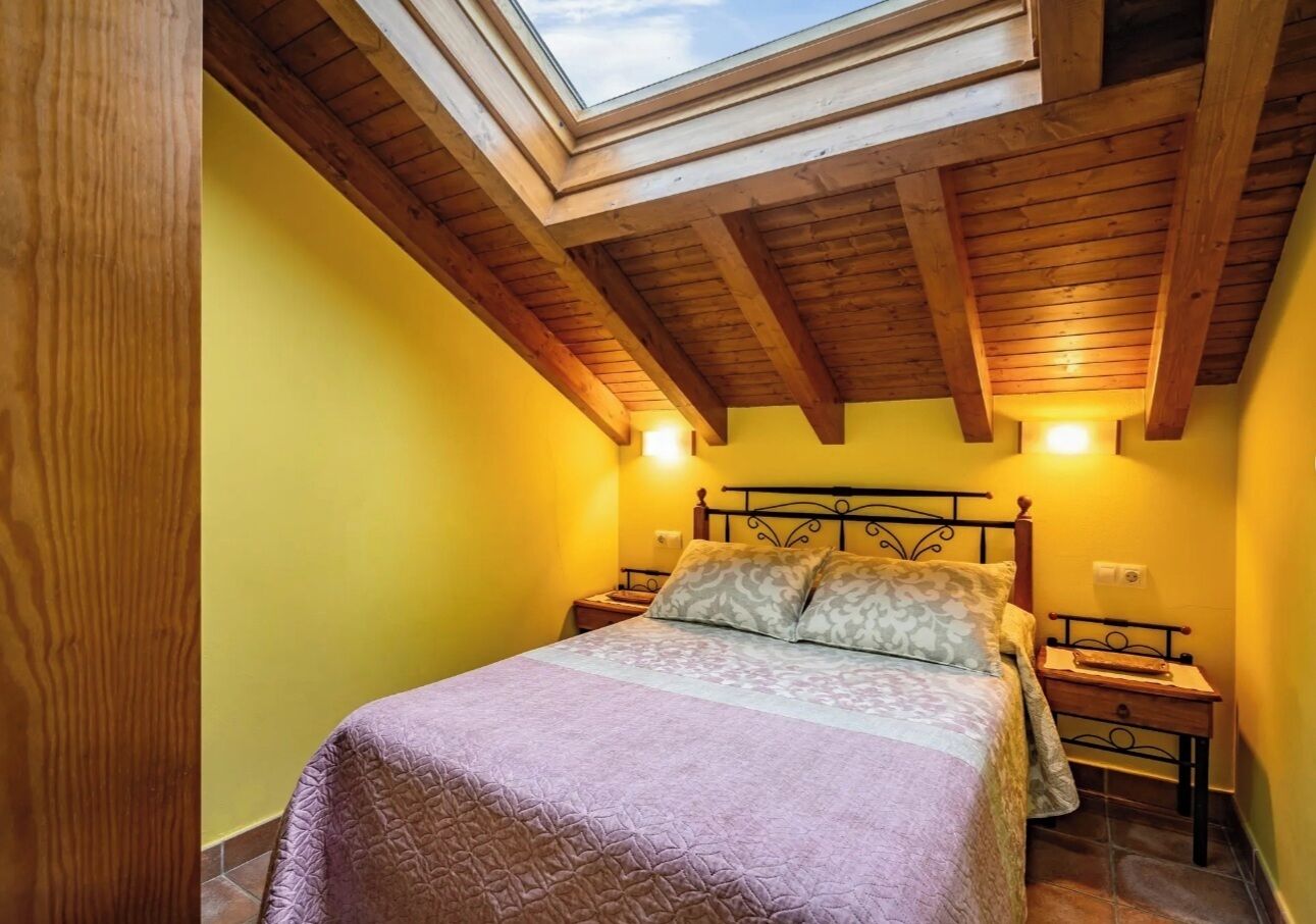 A bedroom with a skylight, wooden beams, a bed with patterned bedding, and bedside tables.