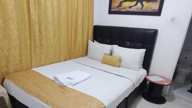 Un lit bien fait, recouvert d’une couvre-lit blanche et dorée, avec une tête de lit noire et une petite table de chevet dont le plateau est rouge.