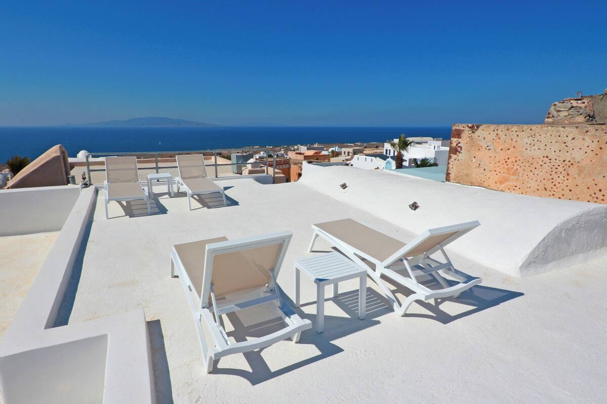 Eine Dachterrasse mit weißen Liegestühlen und Tischen, mit Blick auf das Meer und entfernte Berge.