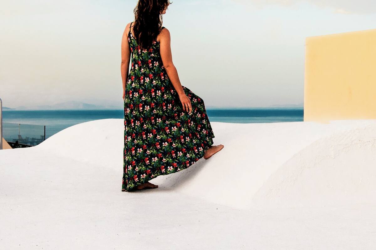 Eine Frau in einem Blumenkleid sitzt auf einer weißen Fläche, in der Nähe befindet sich ein rotes Objekt, mit Blick auf ein Gewässer und einen klaren Himmel.
