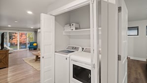 A laundry room with a washer and dryer, a white door, and a view of a living room with a blue chair and a TV. - Adventure Awaits at The Lake Loft (Coeur d`Alene)