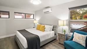 A modern hotel room with a bed, two bedside lamps, a small wooden nightstand, a blue armchair, and a large window with a view of a garage and trees. - Adventure Awaits at The Lake Loft (Coeur d`Alene)