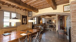 Interior rústico com vigas de madeira aparentes, piso de azulejos e arco de pedra que dá acesso a uma cozinha.