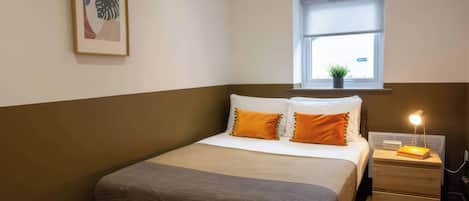 A neatly arranged bedroom with a bed, a bedside table with a lamp, a window with a view, and a framed picture on the wall.