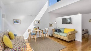 Um espaço de estar moderno estilo loft, com pé direito alto, área de jantar com mesa redonda e cadeiras, sofá com almofadas coloridas e uma grande janela que proporciona luz natural.