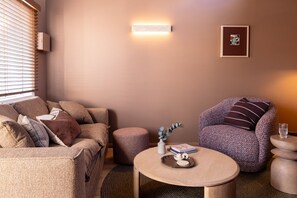Un salon chaleureux avec un canapé, un fauteuil, un pouf et une table basse ronde agrémentée de livres et d’un vase.