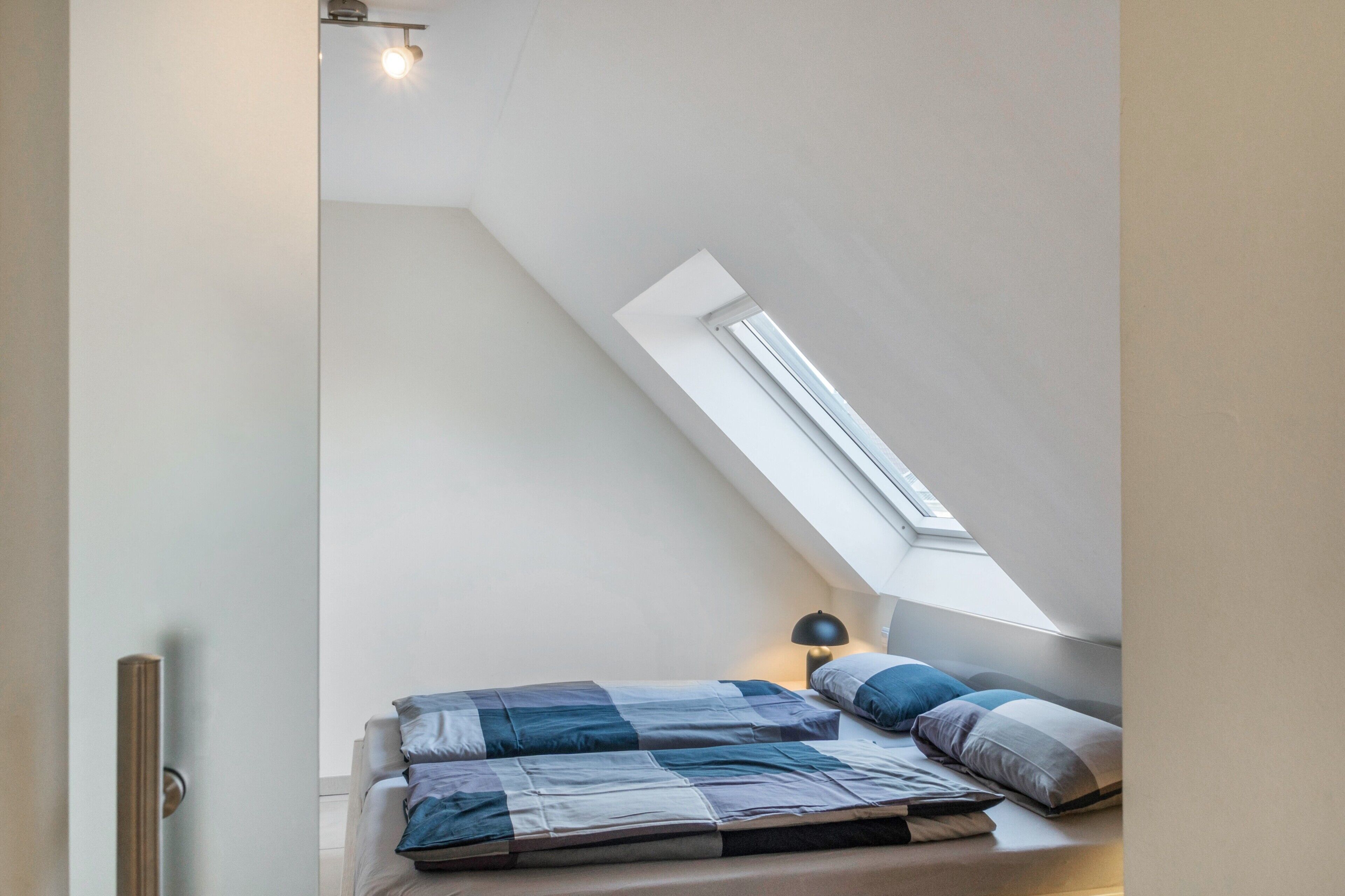 Une chambre avec un plafond en pente, une lucarne, un lit recouvert d’une couette bleue et blanche, et une lampe de chevet.