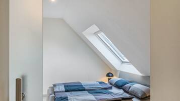 Une chambre avec un plafond en pente, une lucarne, un lit recouvert d’une couette bleue et blanche, et une lampe de chevet.