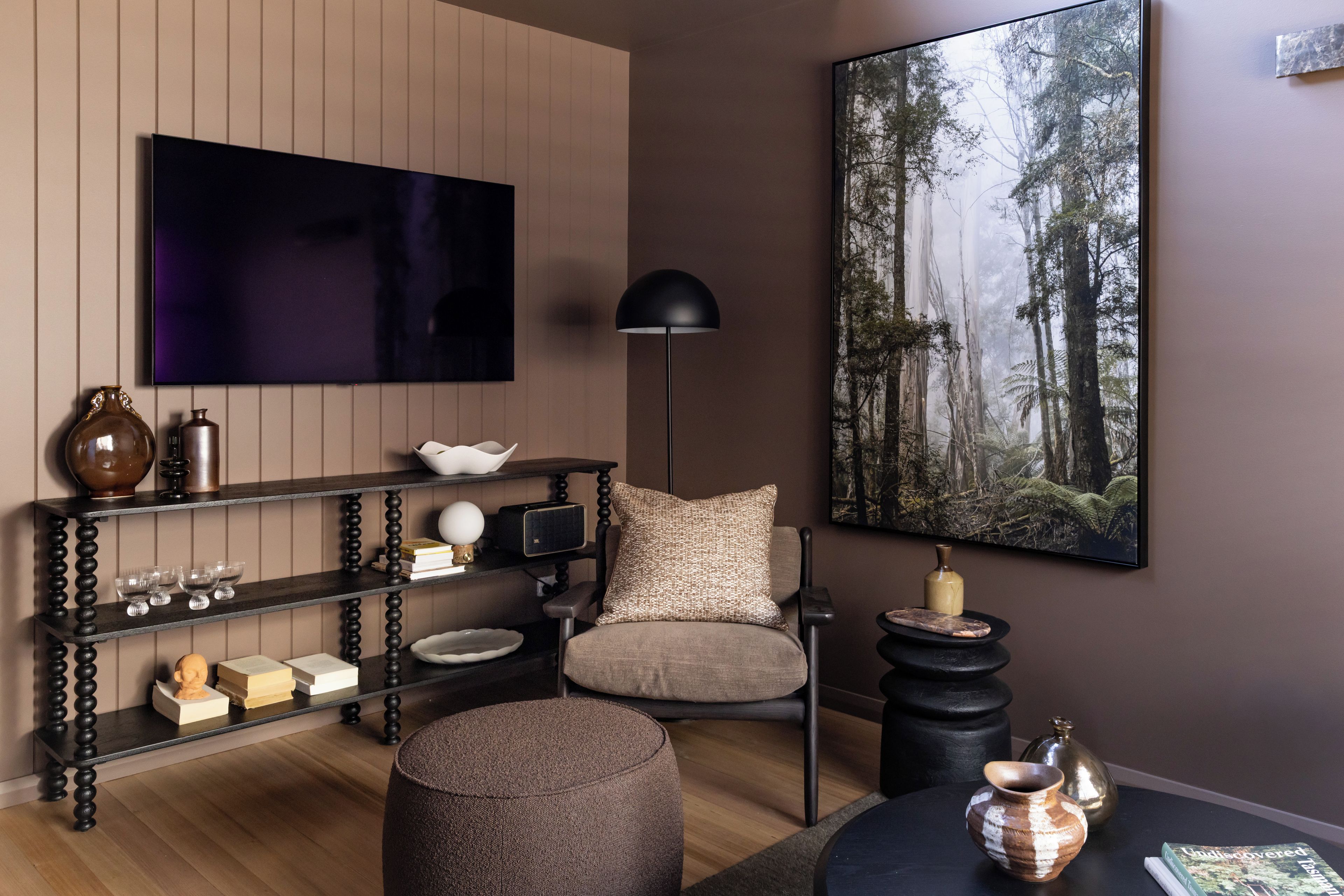 A modern living room with a flat-screen TV, a bookshelf, a round ottoman, and a large forest-themed painting.