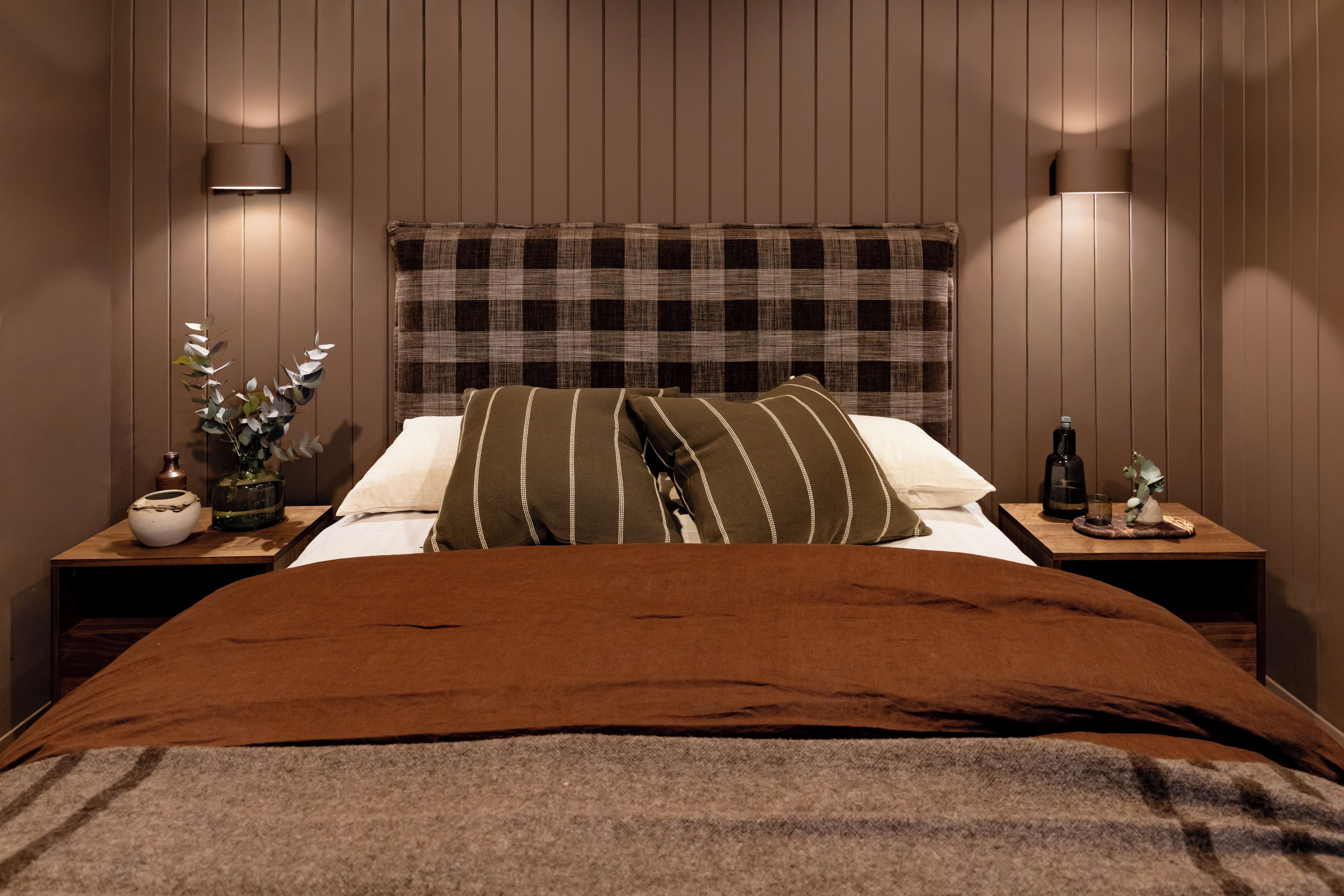 Ein Schlafzimmer mit einem Holzkopfteil, einer braunen Bettdecke, zwei Kissen, einem Nachttisch mit einer Vase und einer Flasche sowie einer Wandlampe.