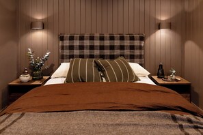 Een slaapkamer met een houten hoofdbord, een bruine spreidekbed, twee kussens, een nachtkastje met een vaas en een fles, en een wandlamp.
