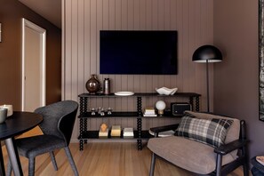 Ein modernes Wohnzimmer mit einem wandmontierten Flachbildfernseher, einem schwarzen Konsolentisch mit Dekorationsobjekten, einem Sofa mit kariertem Kissen und einer runden Stehlampe.