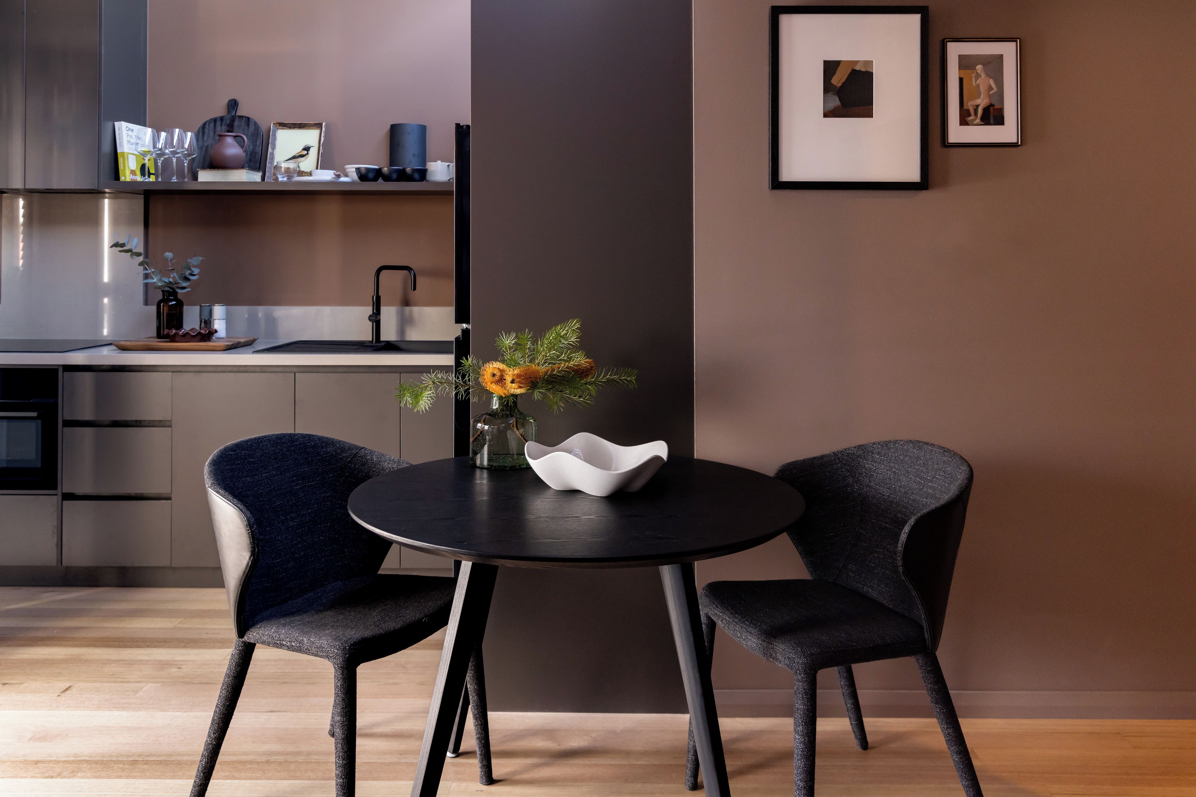 A modern kitchen with a dining area featuring two chairs and a table, a vase with flowers, and framed artwork on the wall.