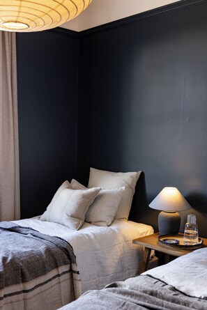 Un lit bien fait, avec des oreillers et une couverture, une table de chevet avec une lampe et un verre d’eau, et un mur sombre.
