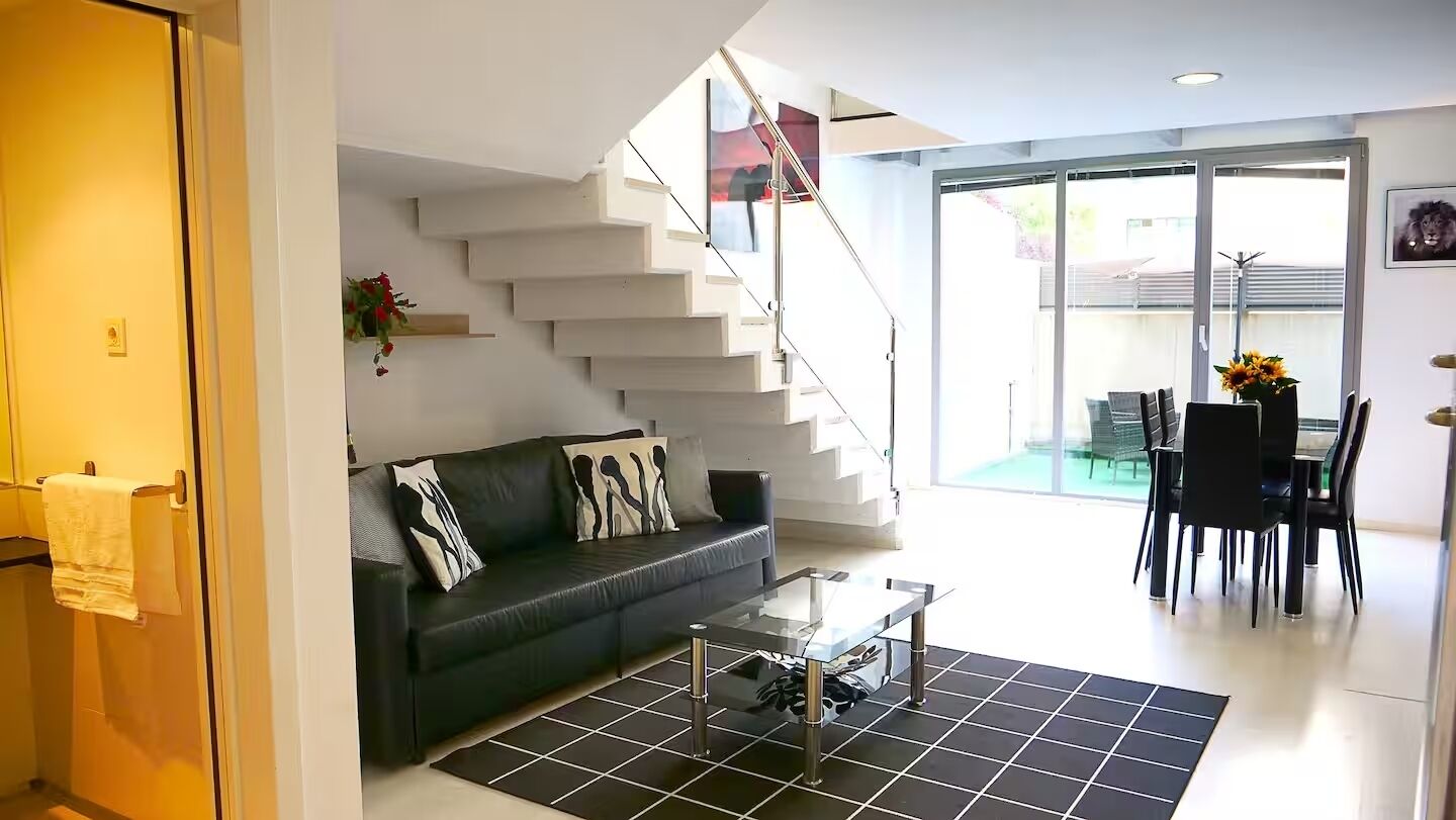 Un salon moderne avec un escalier, un canapé agrémenté de coussins à motifs, une table basse et un coin repas avec des chaises et une table.