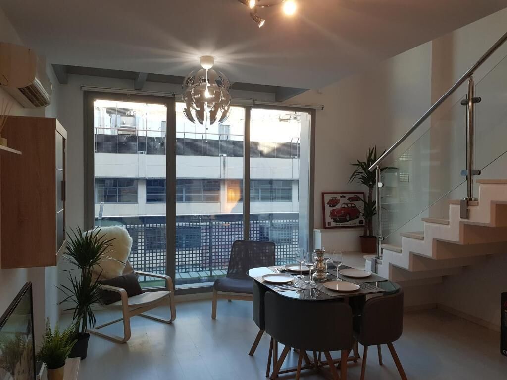 Et moderne spiseområde med et glasbord, stole, en lysekrone og udsigt til en bygning gennem store vinduer.