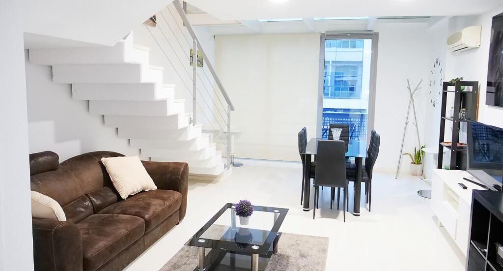 Een moderne woonkamer met een bruine leren bank, een glazen salontafel, een eethoek met een zwarte tafel en stoelen, en een trap naar een verdieping boven.