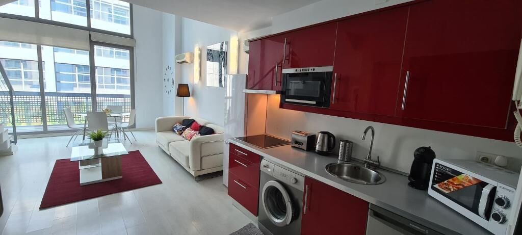 Un apartamento moderno con cocina, que cuenta con gabinetes rojos, horno empotrado y lavadora. Hay una zona de comedor con mesa y sillas, y un sofá en la sala.