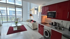 Eine moderne Wohnung mit einer Küche, die rote Schränke, einen Backofen und eine Waschmaschine beinhaltet. Es gibt einen Essbereich mit Tisch und Stühlen sowie ein Sofa im Wohnzimmer.