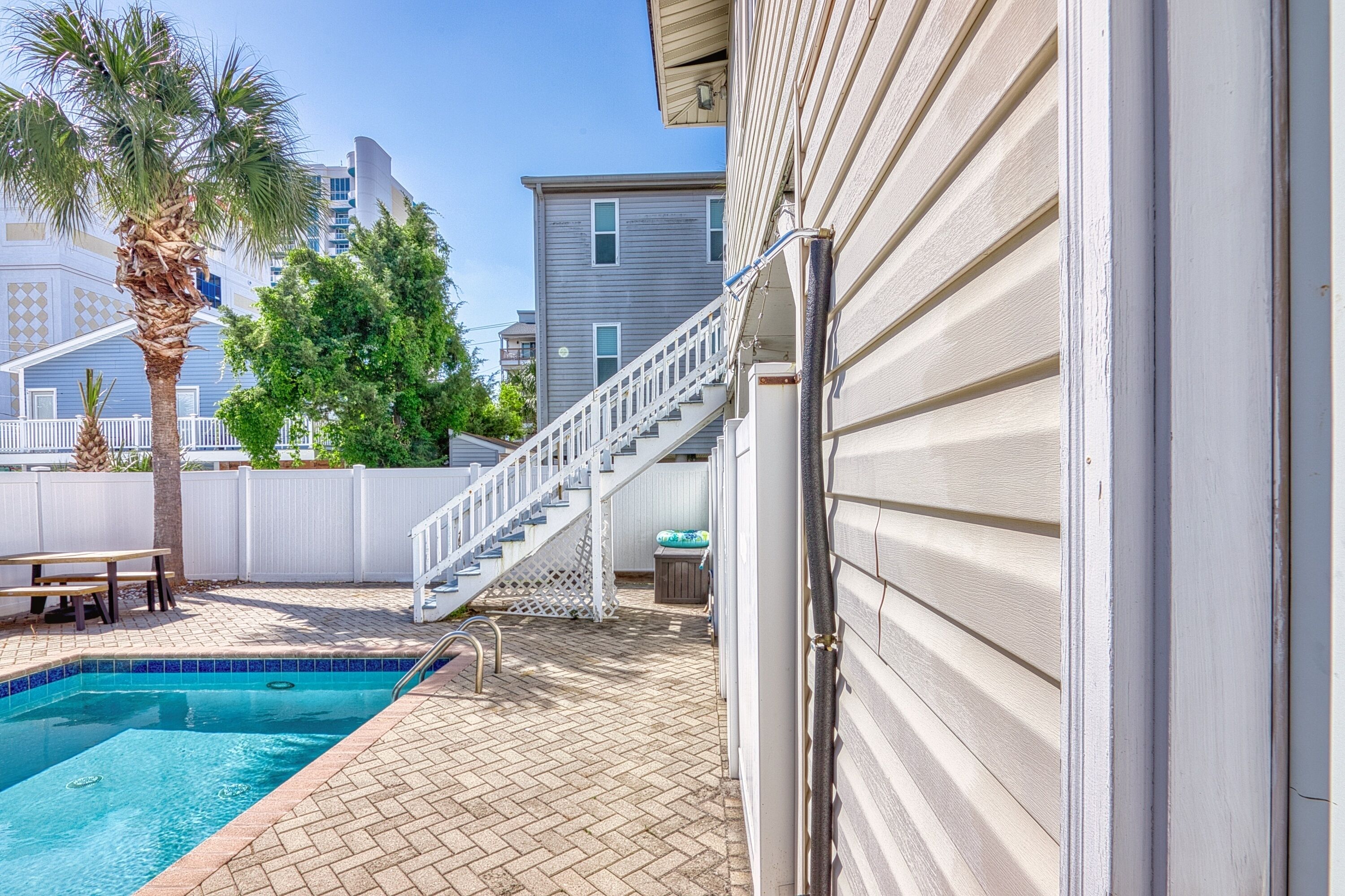 A swimming pool with a wooden deck, a white fence, and a staircase leading to a building.