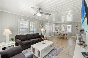 A spacious living room with a dark brown sofa, a white coffee table, and a ceiling fan. There is a dining area with a table and chairs, and a kitchen area with a stove and sink. - Bella Sirena- 3BR/1.5BA Dog Friendly with Pool (North Myrtle Beach)