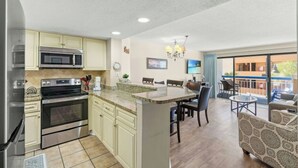 A modern kitchen with built-in appliances, a dining area with a large table and chairs, and a balcony with outdoor seating. - Forest Dunes 204- Side Ocean View 1 Bedroom- Pool (Myrtle Beach)