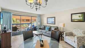A hotel room with a balcony, a sofa, a dining table, and a chandelier. - Forest Dunes 204- Side Ocean View 1 Bedroom- Pool (Myrtle Beach)