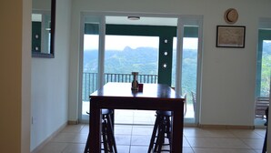 Un comedor con mesa y taburetes, un ventilador de techo, vistas a montañas a través de un ventanal y un cuadro en la pared.