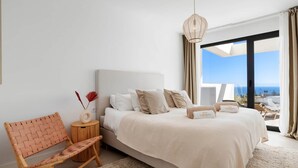 Quarto com cama, cadeira, mesa de cabeceira e vista para o mar.
