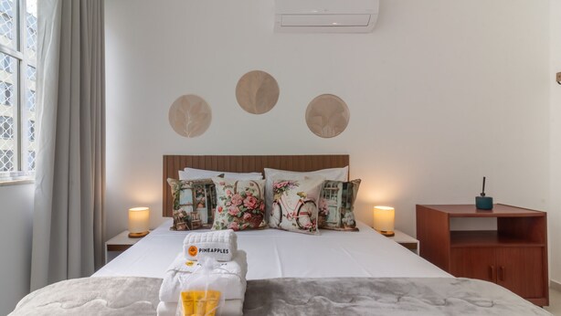 Een hotelkamer met een net opgemaakt bed, bloemenkussens, een houten hoofdbord, een nachtkastje met een lamp en een klein houten kastje.