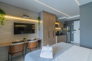 Una habitación de hotel moderna con una televisión de pantalla plana montada en una pared de ladrillo, un escritorio de madera con dos sillas, una kitchenette con refrigerador y una cama con colcha blanca y toallas.