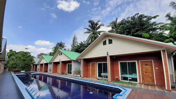 A swimming pool with a tiled deck, adjacent to a row of single-story buildings with large windows and a red roof. - Phenwaree Amphawa Resort by Zuzu (Samut Songkhram)
