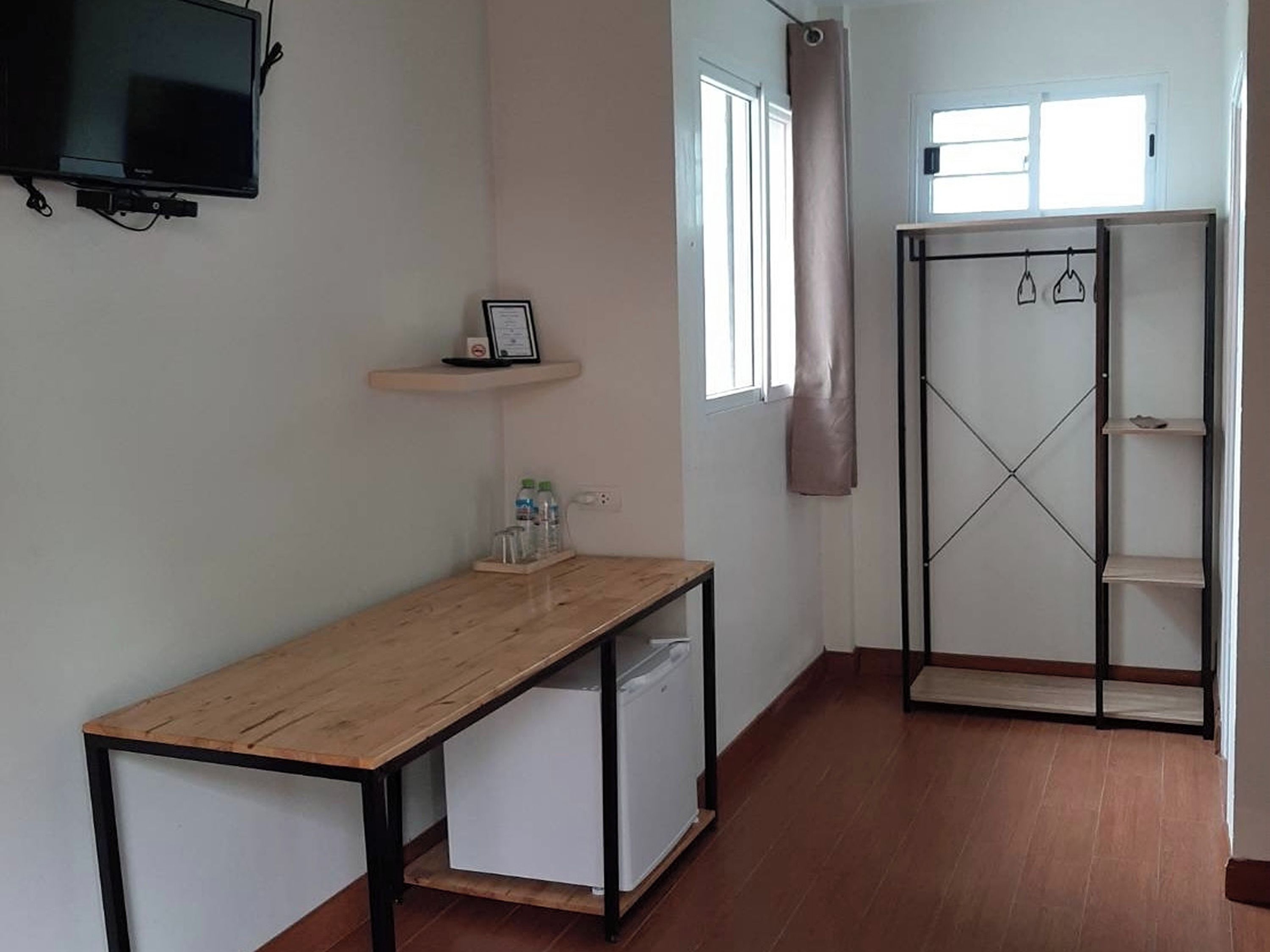 A room with a wooden desk, a white cabinet, a TV mounted on the wall, a window with a curtain, and a wardrobe with shelves.