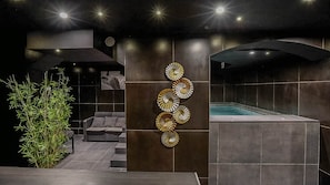 A modern spa area with a hot tub, a wall-mounted decorative panel, and a seating area with a sofa and a plant. - Love Room—Jacuzzi Area / Massage Table (L'Escarène)