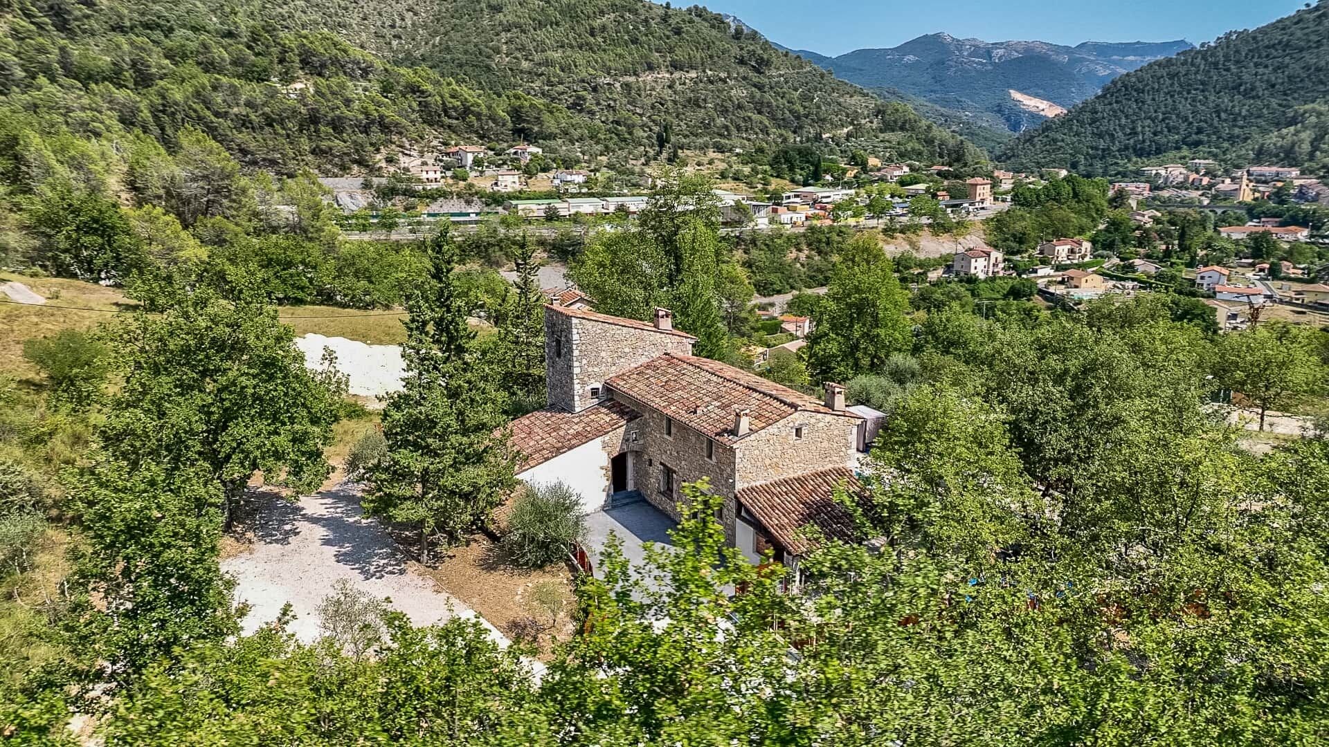 A stone building with a courtyard, surrounded by trees and set against a backdrop of hills and a village.