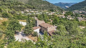 A stone building with a courtyard, surrounded by trees and set against a backdrop of hills and a village. - Love Room—Jacuzzi Area / Massage Table (L'Escarène)